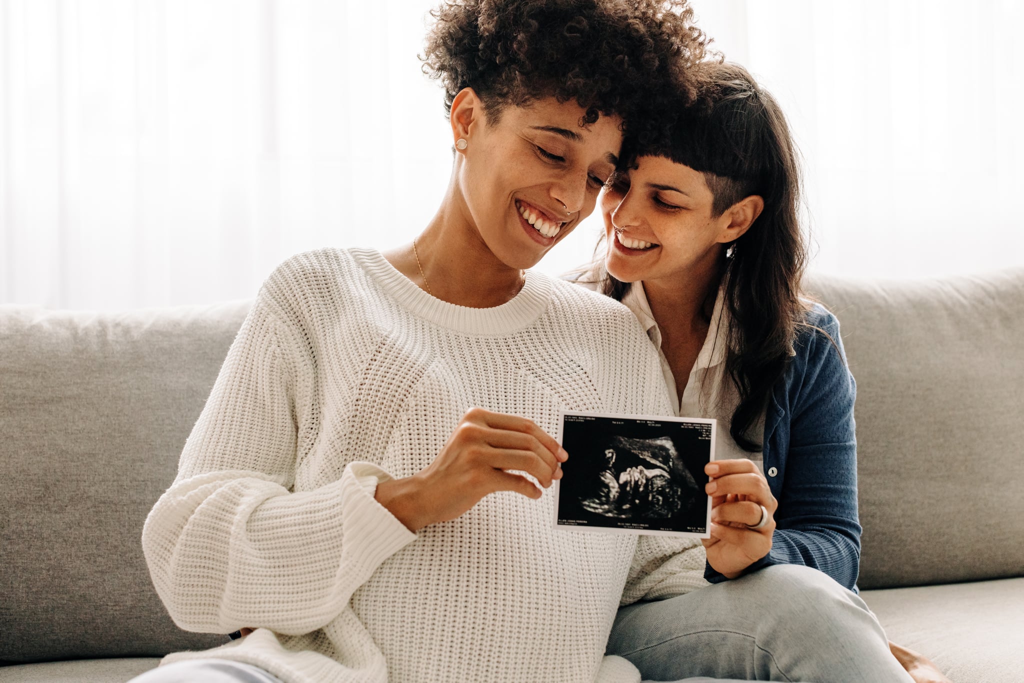 Same sex pregnant couple holding up their ultrasound scan.  Expectant lesbian couple smiling happily while holding an ultrasound scan of their unborn baby.  Young queer couple expecting a baby.