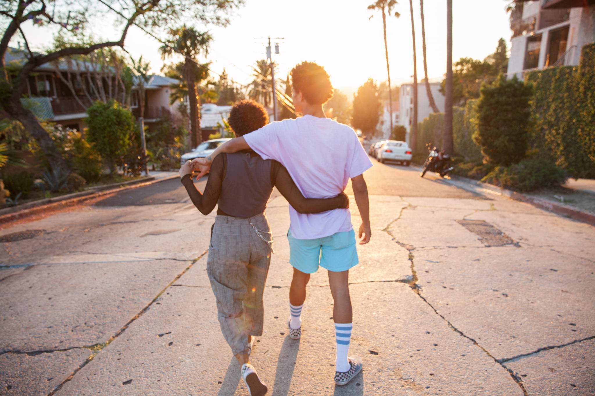 A young African American couple with their arms around each other walking down a suburban street at sunset, shot from behind