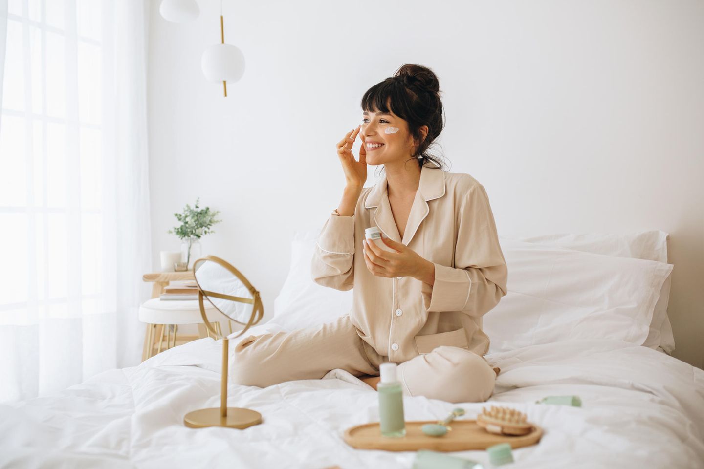 Brunette woman sitting on the bed in pajamas and applying day cream with SPF on her face.